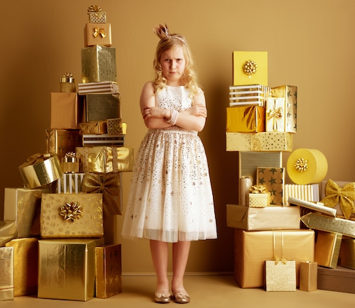 Full length portrait of furious young girl in beige fit and flare dress and a little crown on head standing among 2 piles of golden gifts in front of a plain wall. luxury gifts for demanding customer